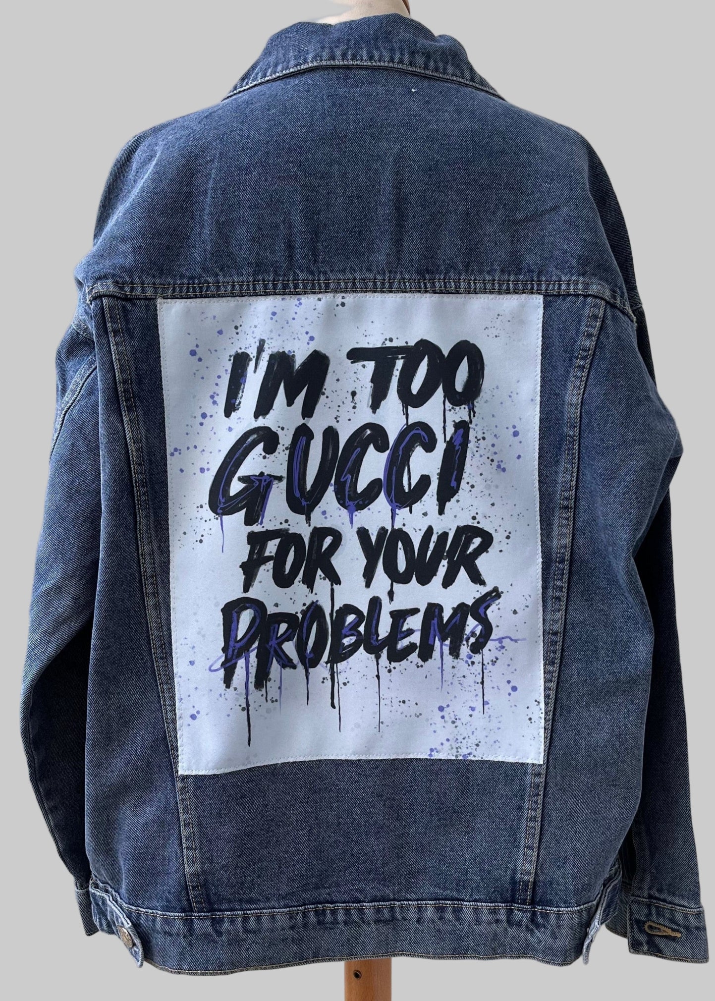 Denim jacket with 'I'm too Gucci for your problems' text on a wooden stand against a light wooden panel background