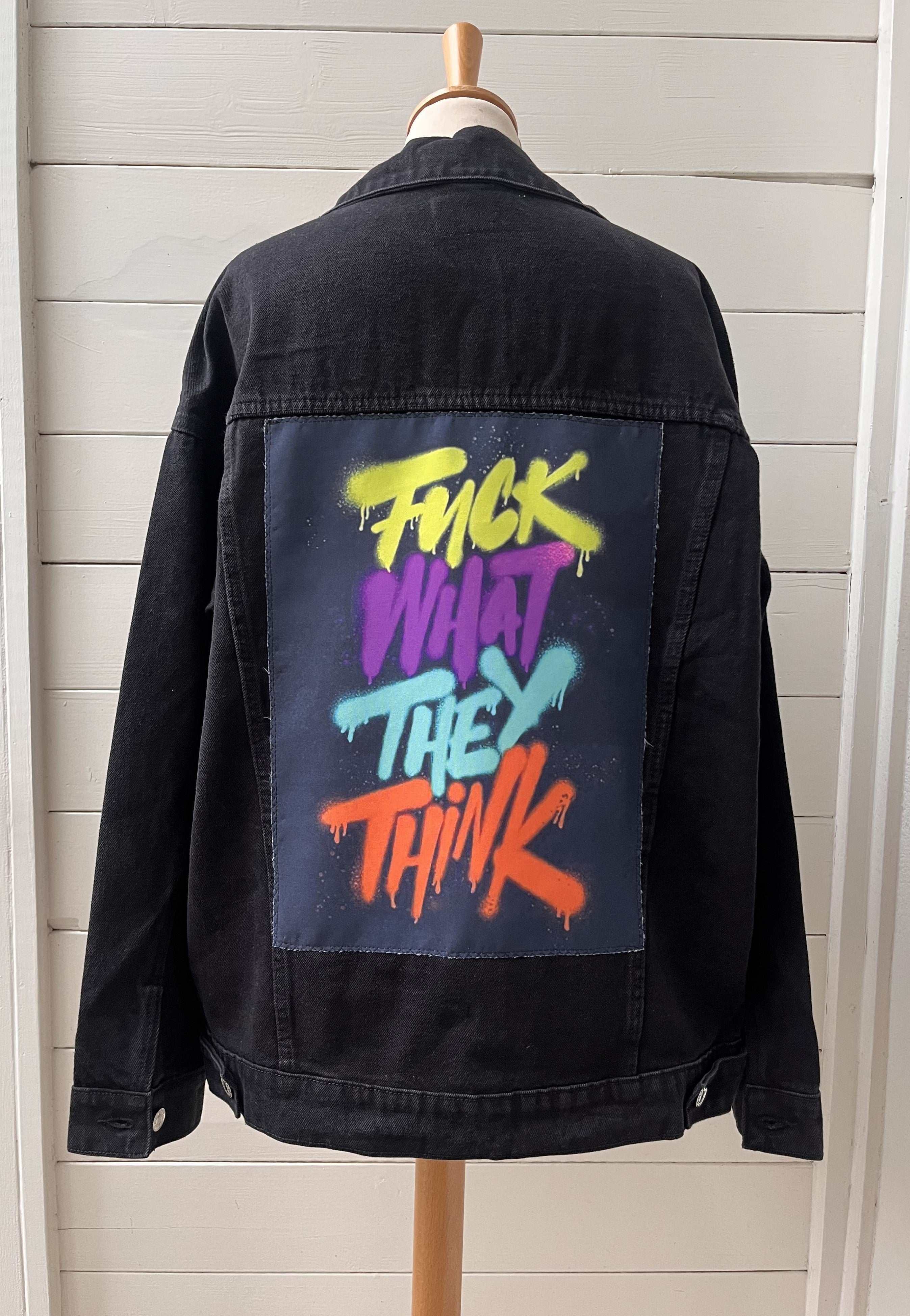 Black denim jacket with colorful text on the back, displayed on a mannequin against a light wooden panel background.