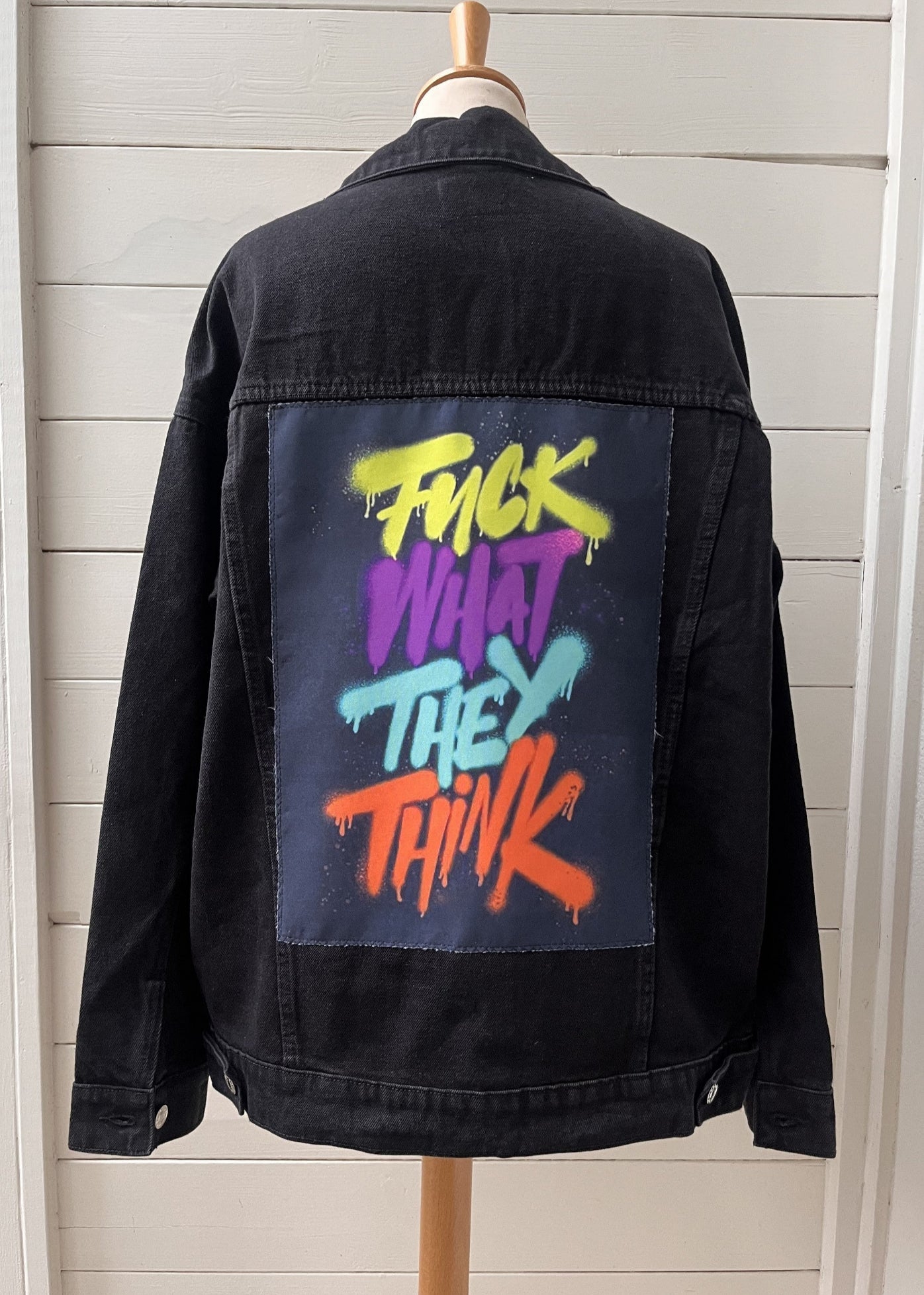 Black denim jacket with colorful text on the back, displayed on a mannequin against a light wooden panel background.
