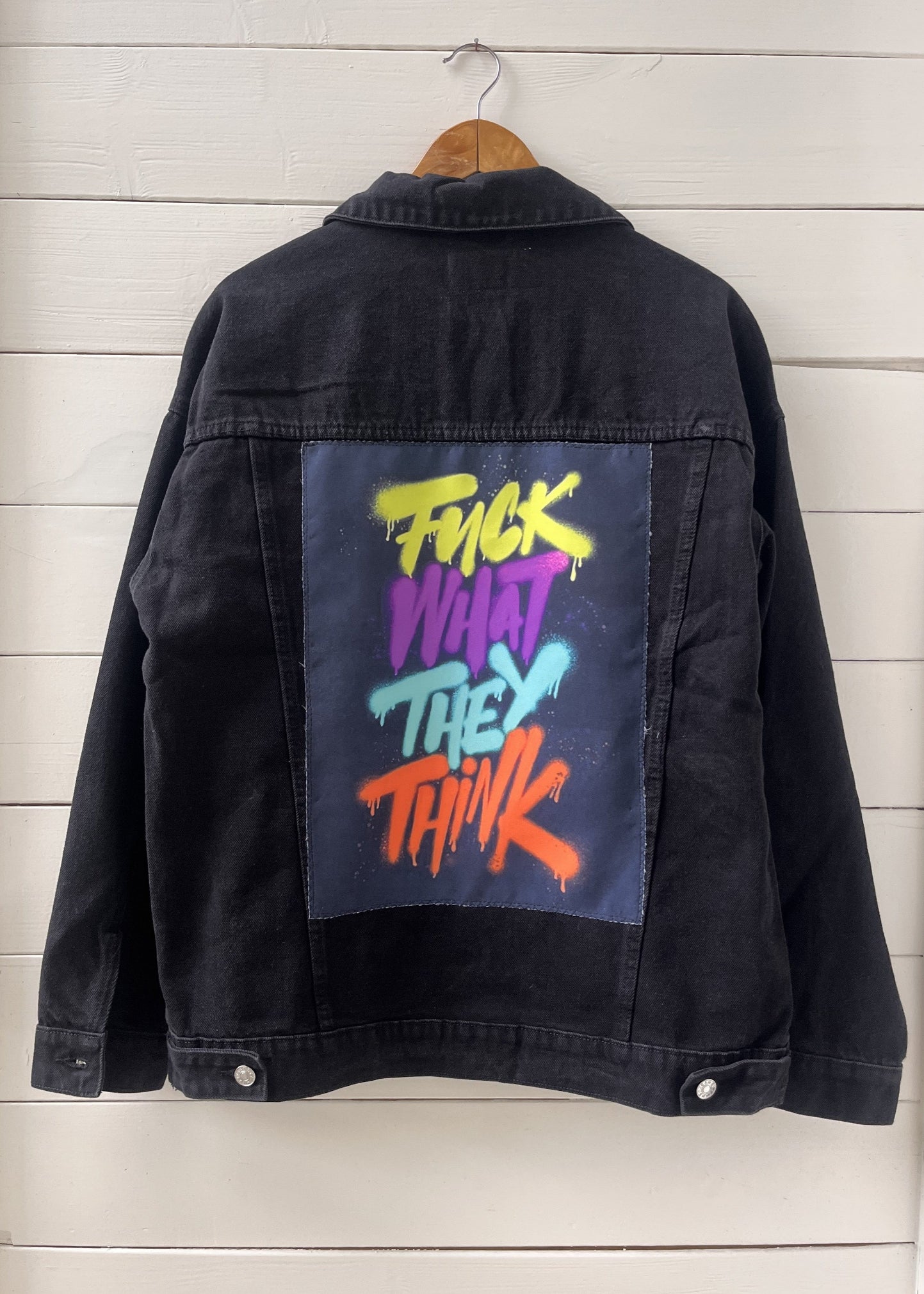 Black denim jacket with colorful text on the back, hanging against a wooden panel background.