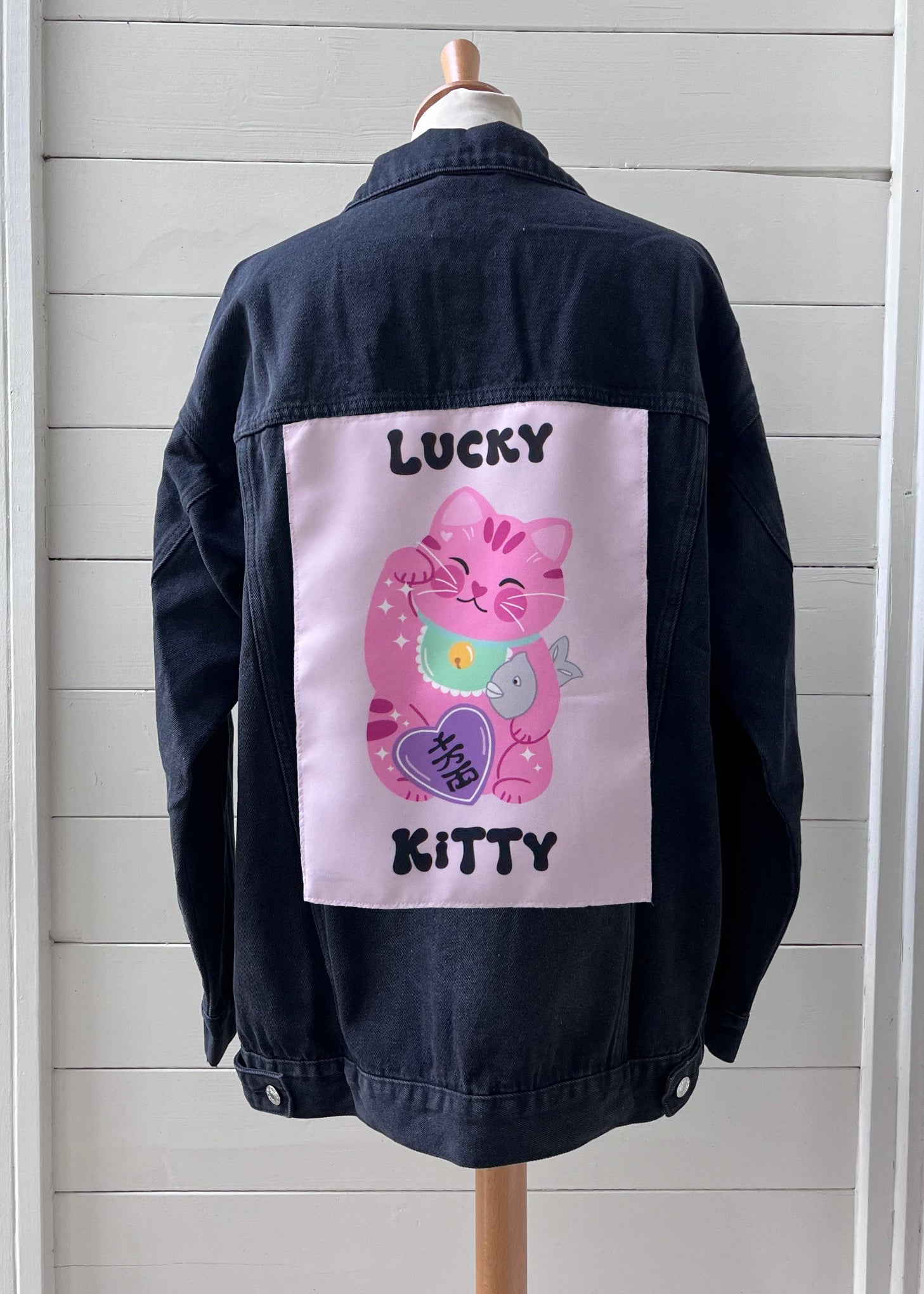 Black denim jacket with a pink cat graphic and text on a wooden mannequin against a light gray background