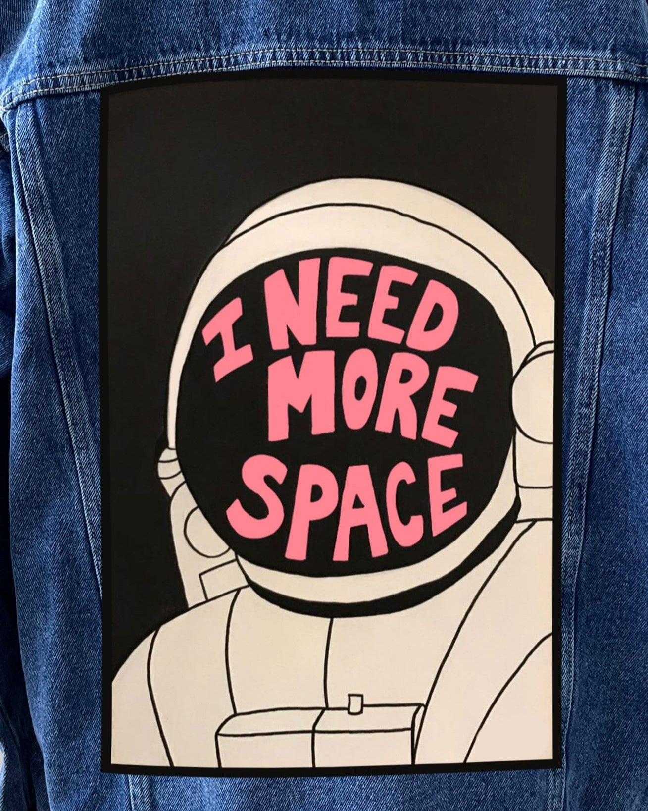 A patch with a cartoon illustration of an astronaut wearing a helmet with the text 'I NEED MORE SPACE' written inside it, attached to a denim fabric.