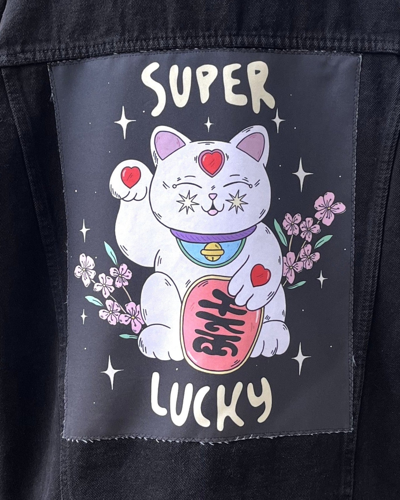 A back patch for a denim jacket featuring an illustration of a white cat with closed eyes, surrounded by floral elements and the text 'SUPER LUCKY'. The patch is black with a printed design.