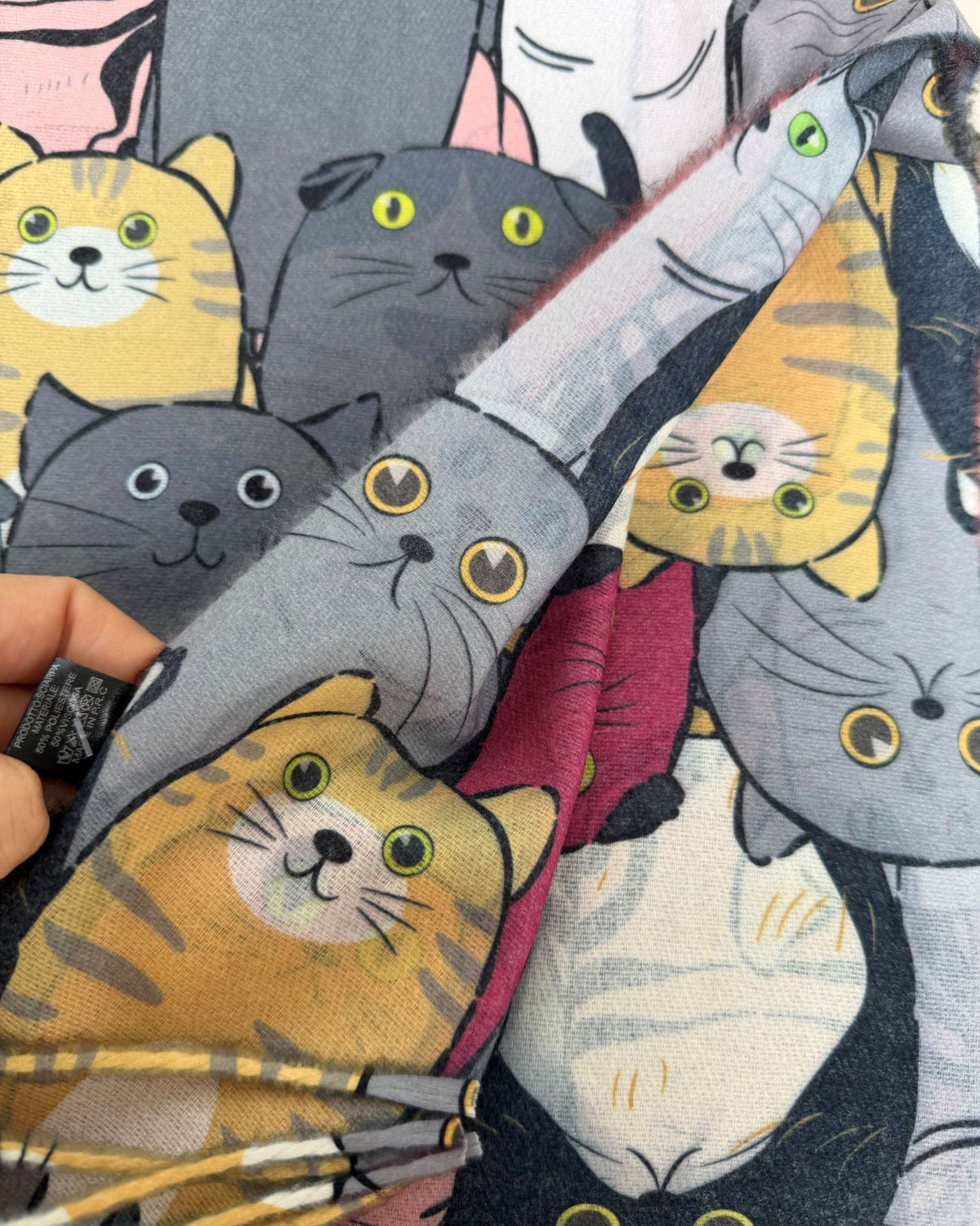 A close-up of a printed fabric featuring cartoon cat faces in gray, yellow, black, white, and pink, with a hand holding one corner to show the material and fringe.