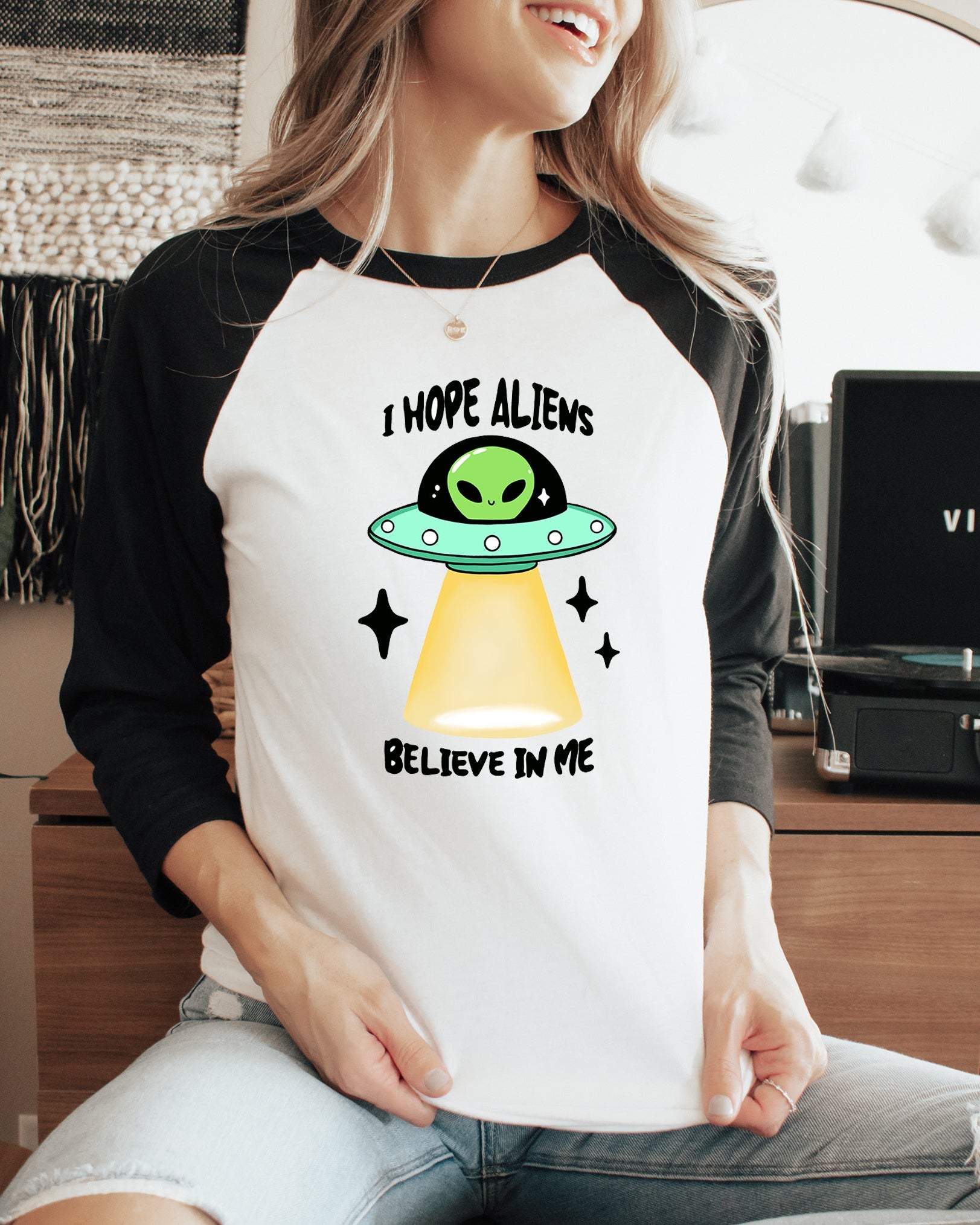 Person wearing a t-shirt with an alien graphic and text, sitting in a room with a plant and record player.