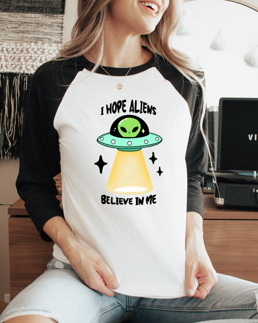 Person wearing a t-shirt with an alien graphic and text, sitting in a room with a plant and record player.