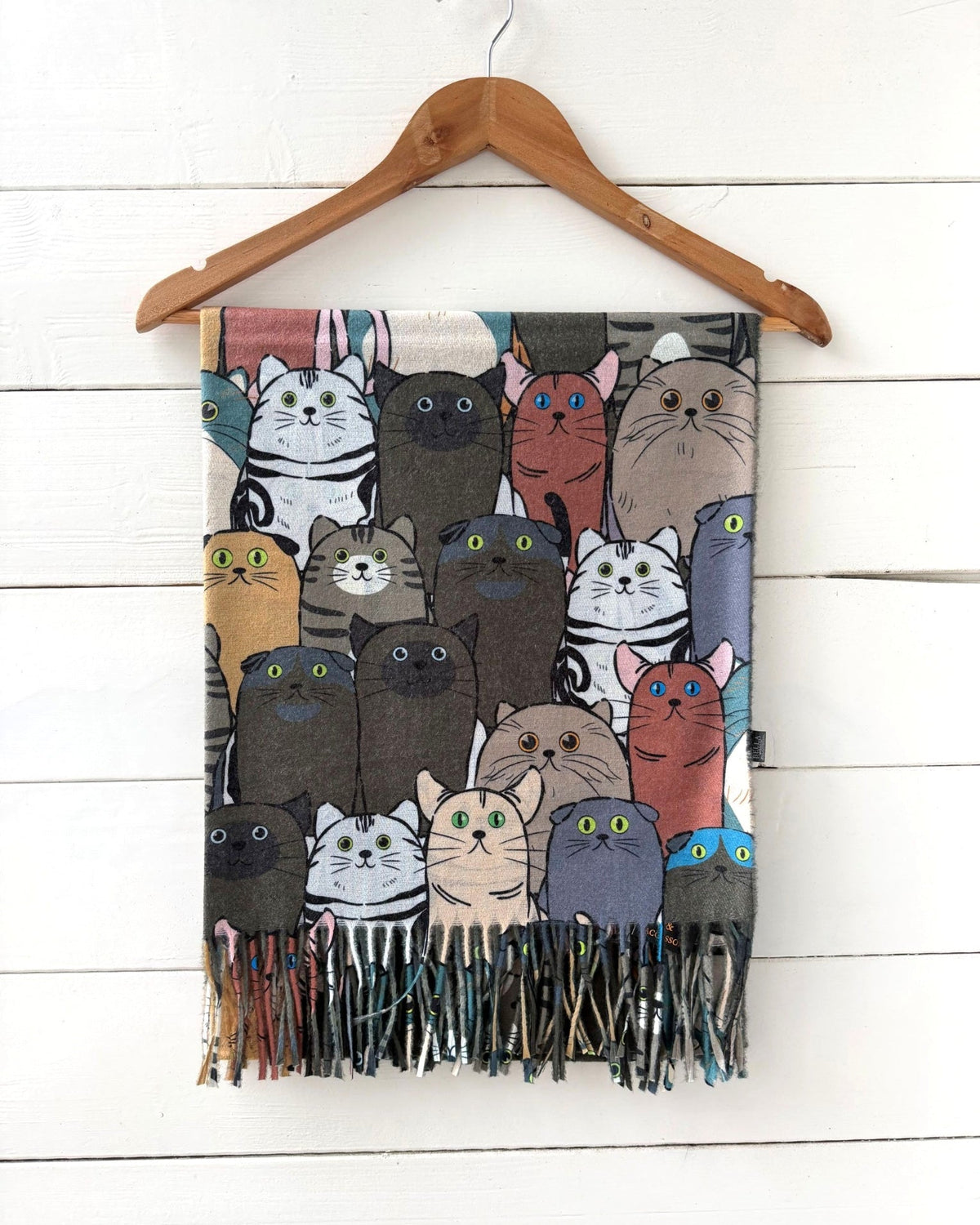 A fringed scarf featuring a colorful cartoon cat pattern, hanging from a wooden hanger against a white wood plank wall.