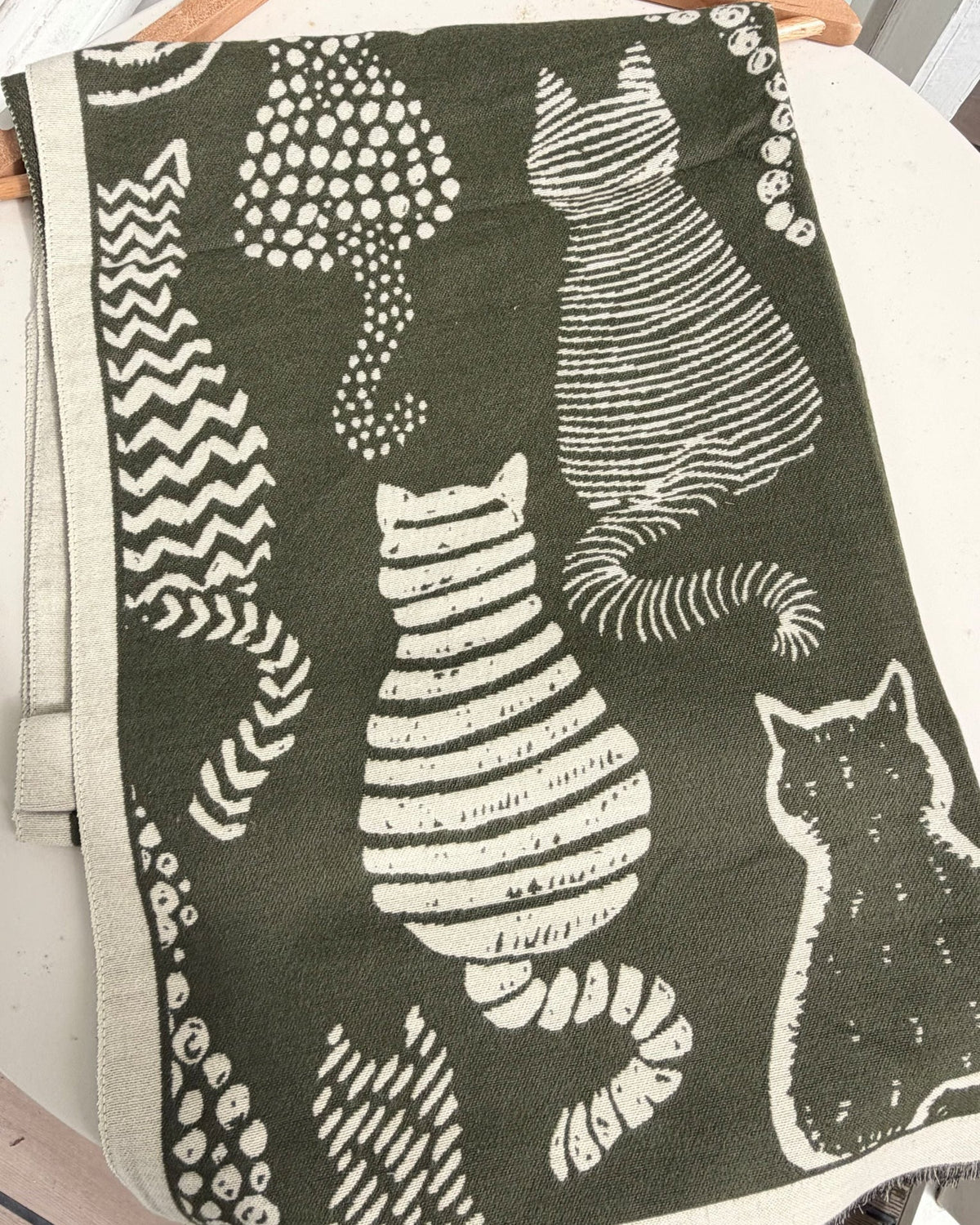 A dark olive green throw blanket featuring a white hand-drawn style print of various cats, draped over a light-colored chair.