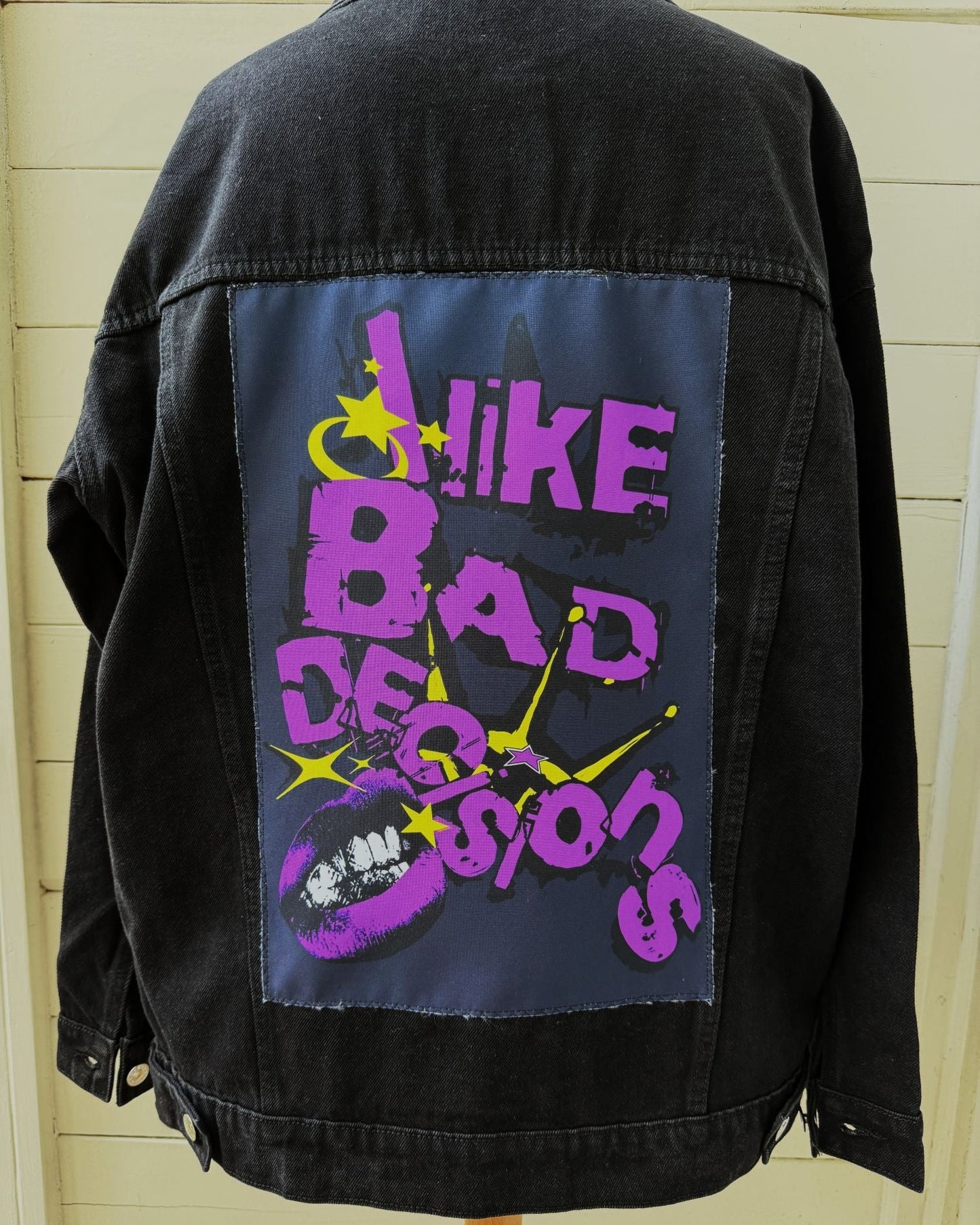 A black denim jacket displayed on a wooden hanger, showing a large back patch with the text 'LIKE BAD DECISIONS' in purple and yellow with a lip graphic.