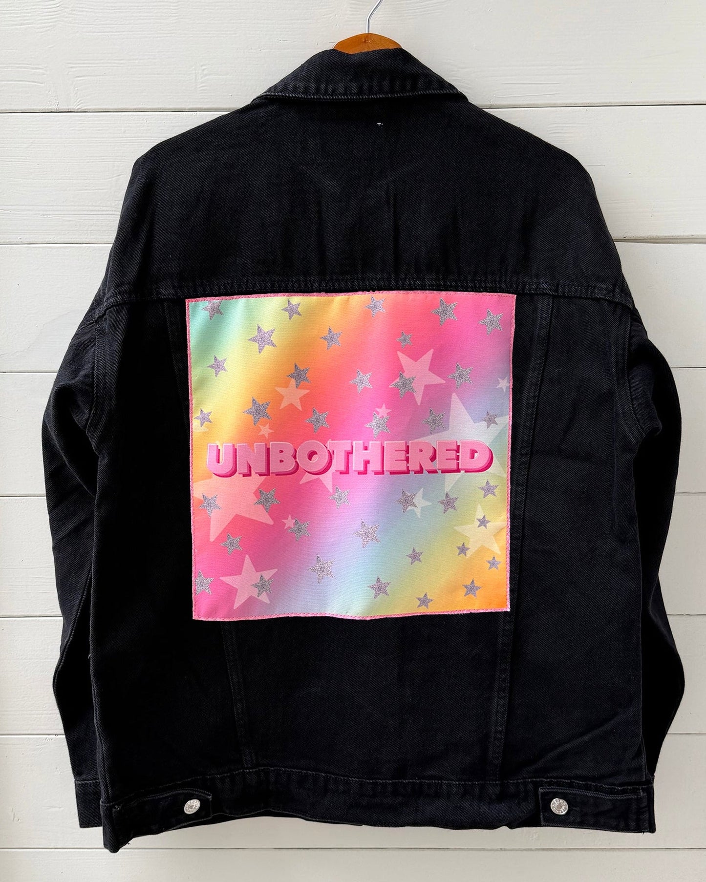Black denim jacket with a colorful 'UNBOtherED' design on a white wooden background