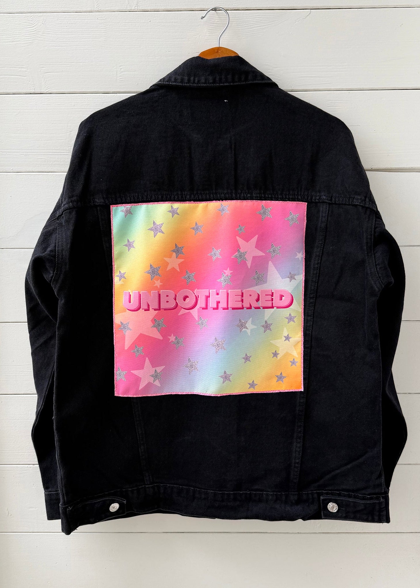 Black denim jacket with a colorful 'UNBOtherED' design on a white wooden background