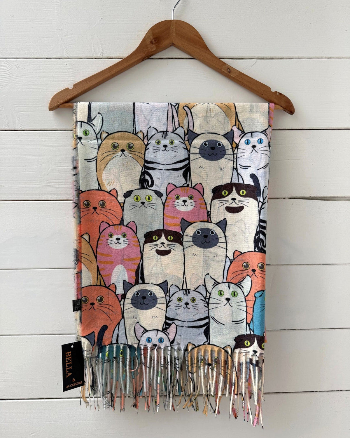 A patterned scarf with colorful cartoon cat illustrations and fringed ends, hanging from a wooden hanger against a white wood plank wall.