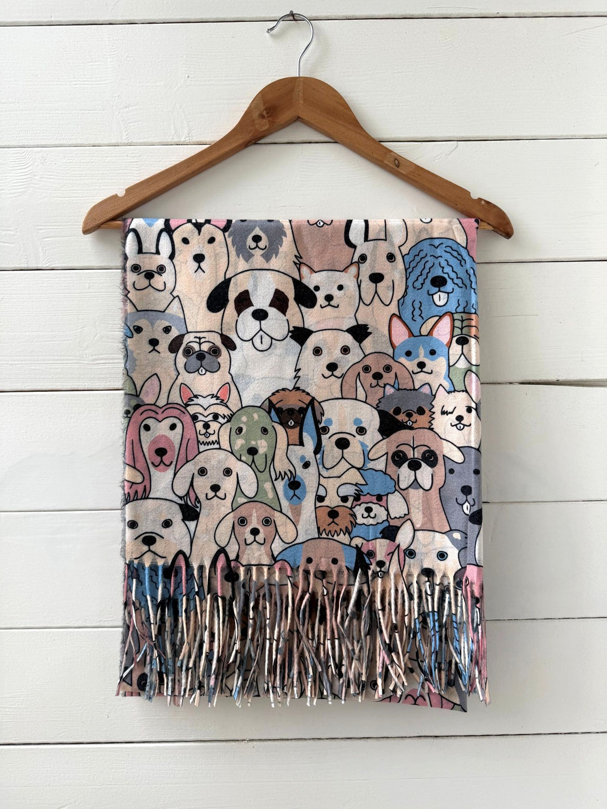 A lightweight scarf featuring a dense cartoon illustration of various dog breeds in pastel colors, hanging from a wooden hanger against a white wood-paneled wall.