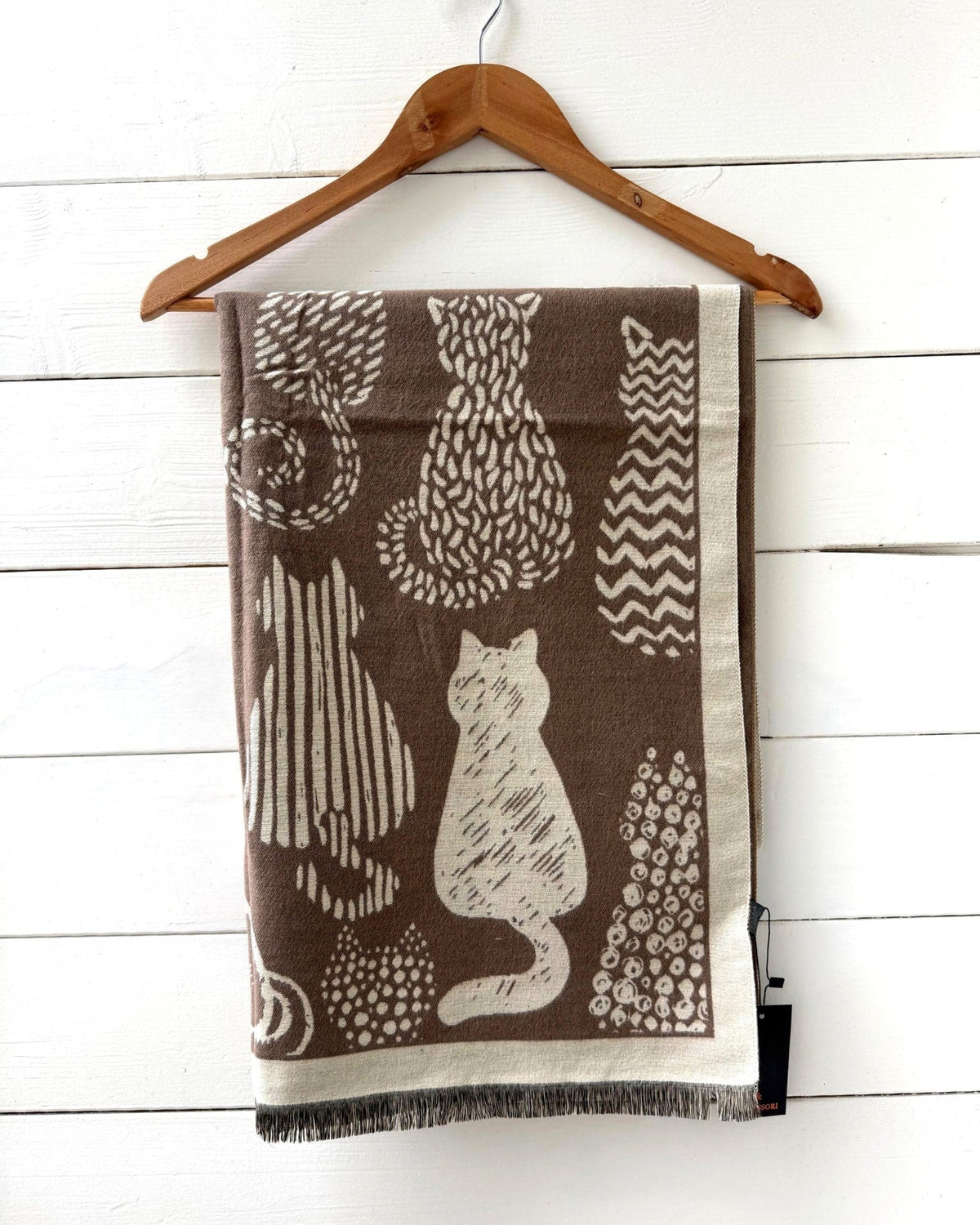 A brown and beige patterned throw blanket with a cat and abstract design, hanging on a wooden hanger against a white wood paneled wall.