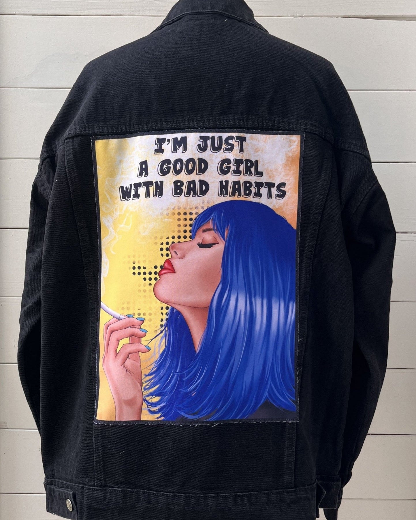 Black jacket with a graphic design of a woman smoking and text on the back, displayed against a light wooden panel background.