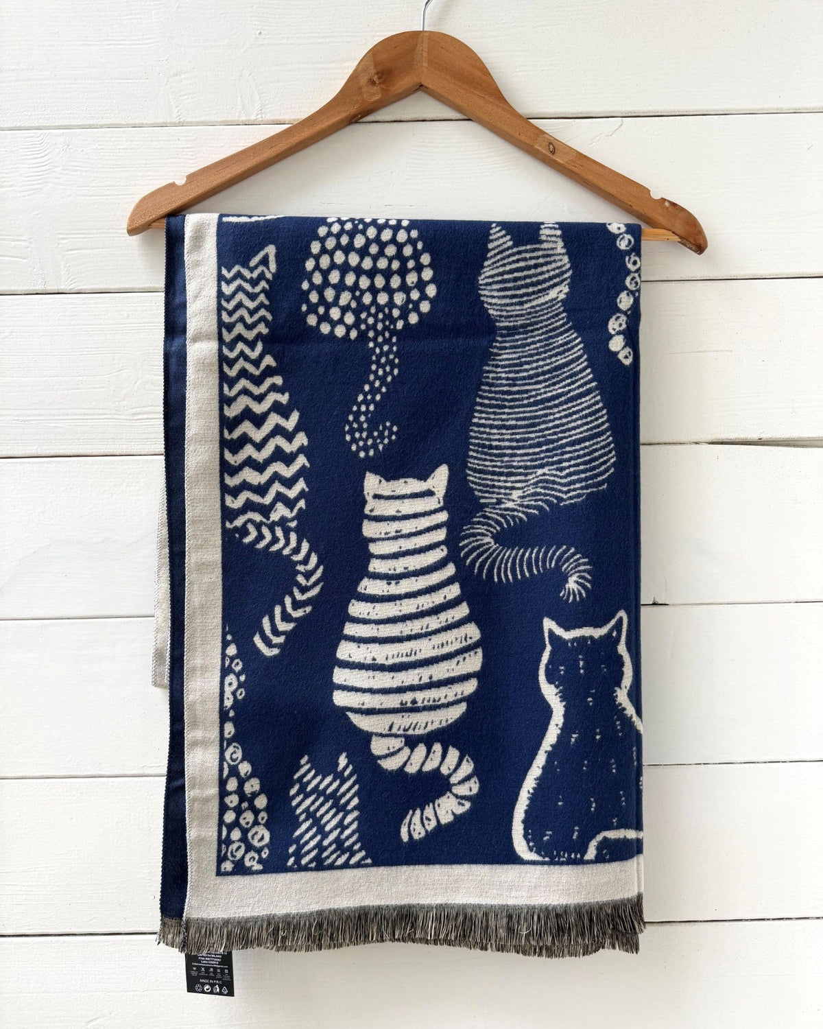 A navy blue scarf with white cat illustrations and fringe, hanging from a wooden hanger against a white wood-paneled wall.