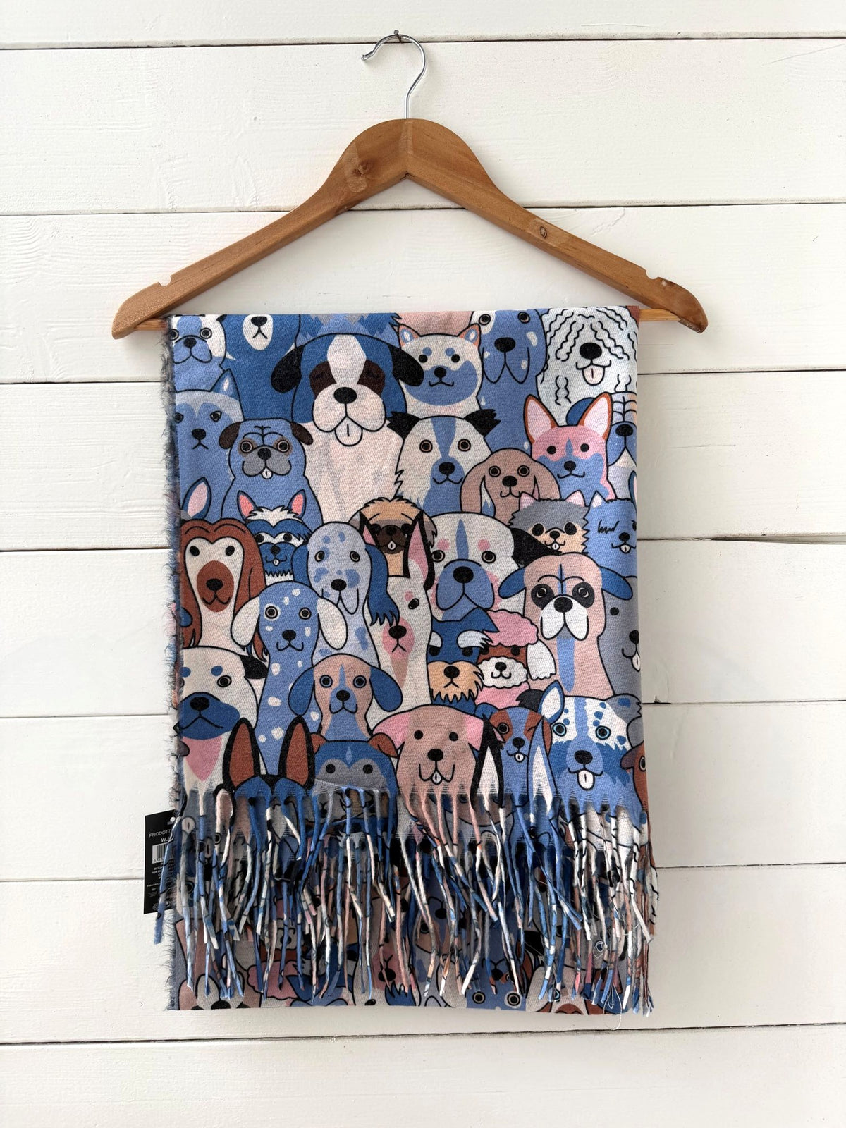 A fringed throw blanket featuring a colorful cartoon dog pattern, hanging from a wooden hanger against a white wood plank wall.