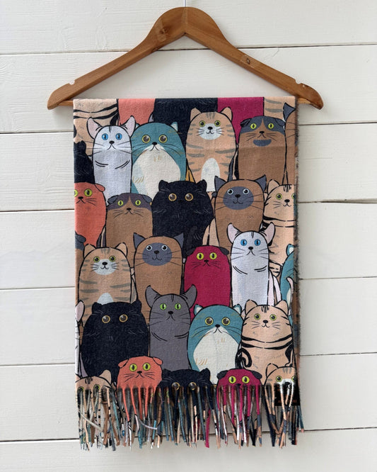 A woven scarf featuring a colorful cartoon cat pattern, hanging from a wooden hanger against a white wooden plank wall.