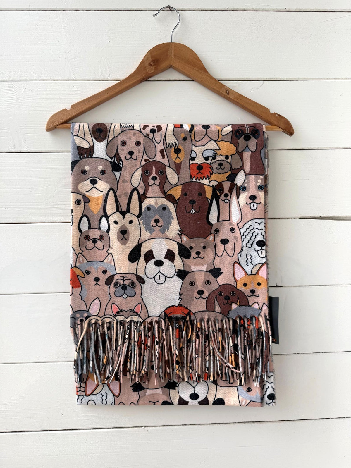 A fringed scarf featuring a dense cartoon illustration of various dog faces, hanging from a wooden hanger against a white wood-paneled wall.