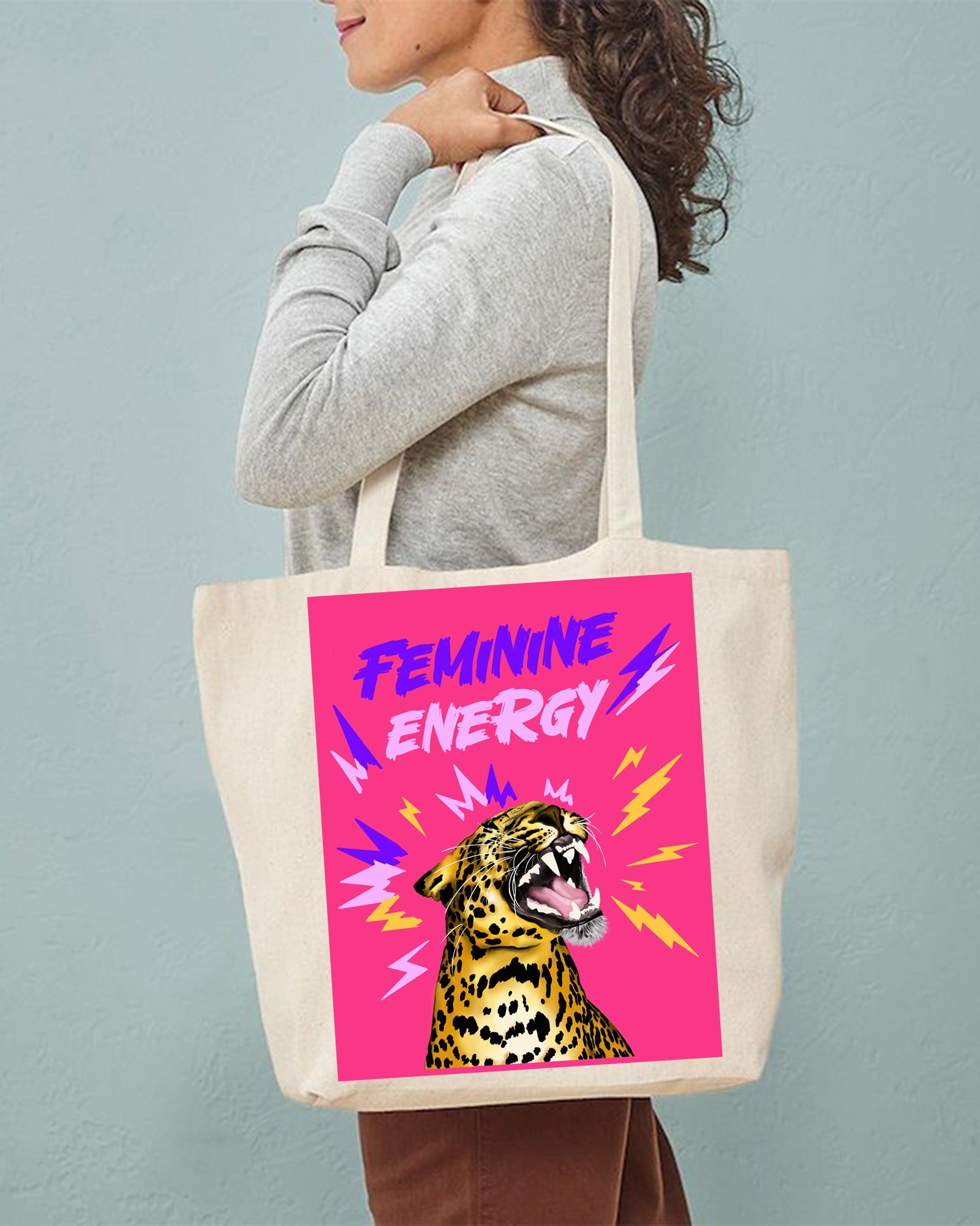Person holding a tote bag with a leopard and 'Feminine Energy' design on a pink background.