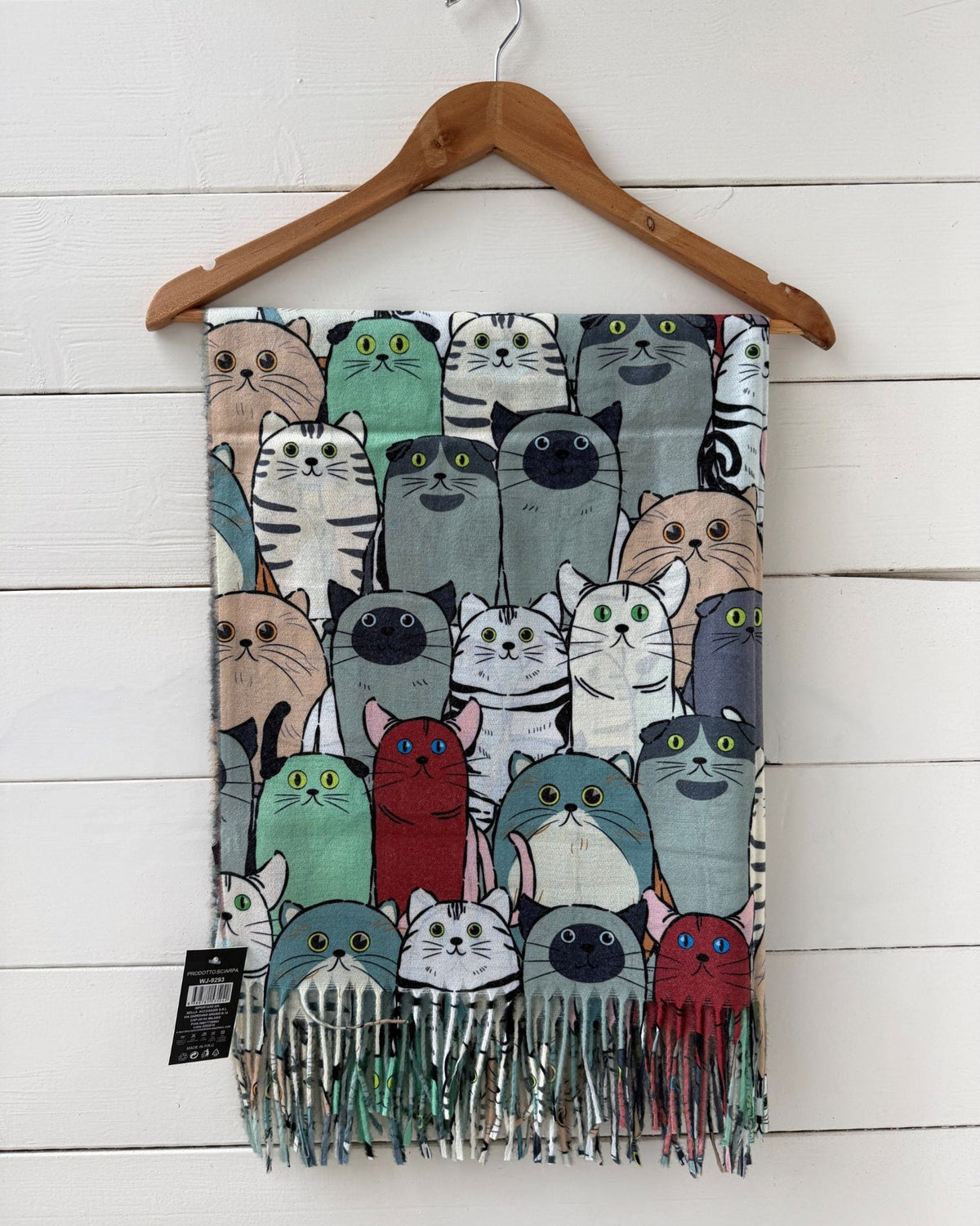 A patterned scarf featuring cartoon cats in various colors, hanging from a wooden hanger against a white wood-paneled wall.