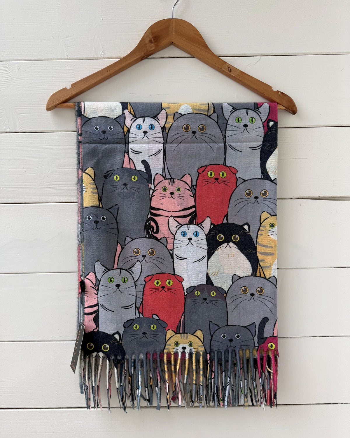 A fringed scarf featuring a dense cartoon cat pattern in various colors, hanging from a wooden hanger against a white wood plank wall.