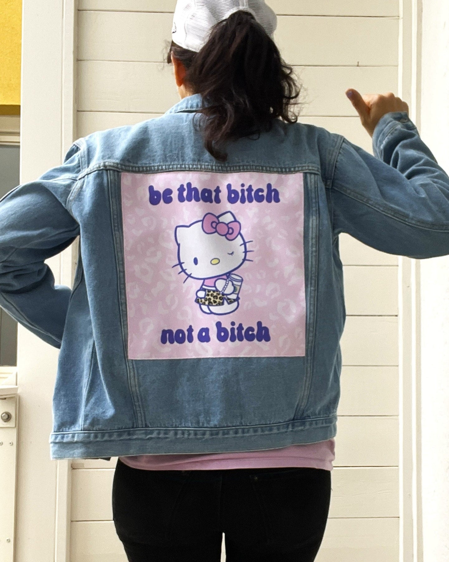 Person wearing a denim jacket with a graphic design and text on the back, standing against a light-colored wall.