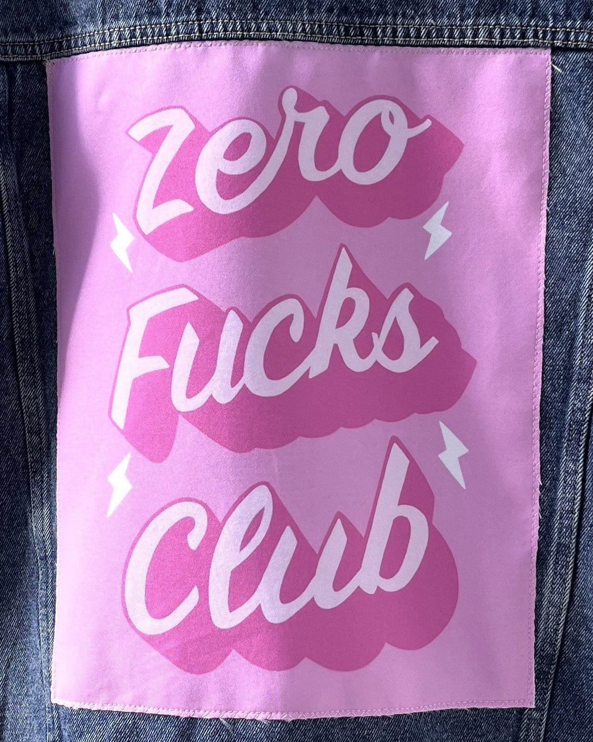 A pink patch with the text 'Zero Fucks Club' printed in a bold white font, attached to a denim fabric.