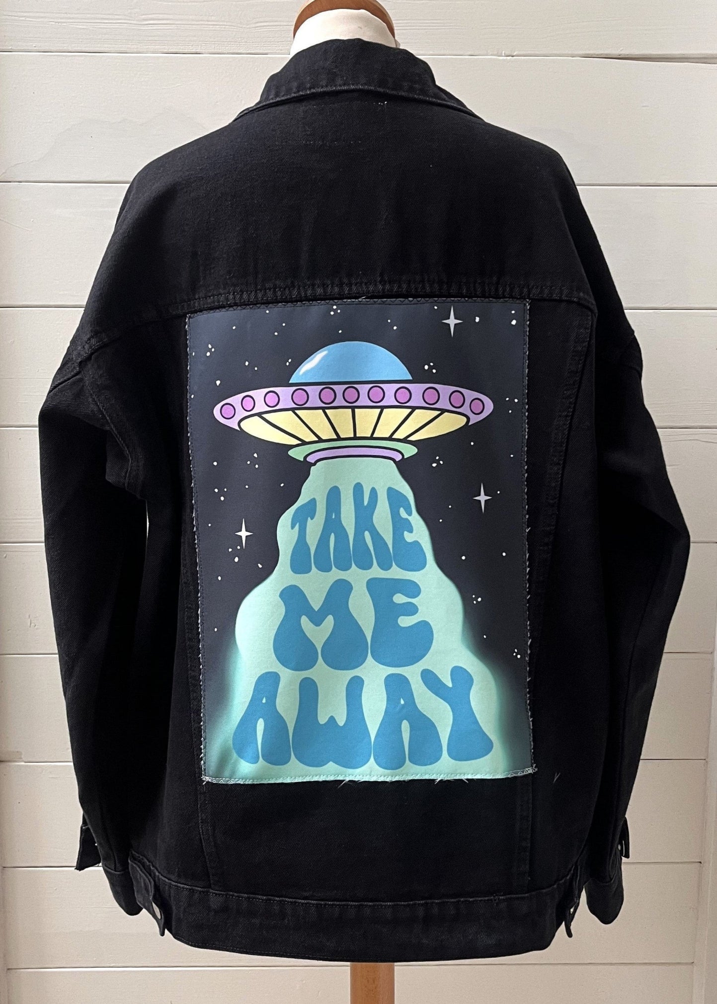 Black jacket with a UFO design and 'Take Me Away' text on the back, displayed against a white wall.