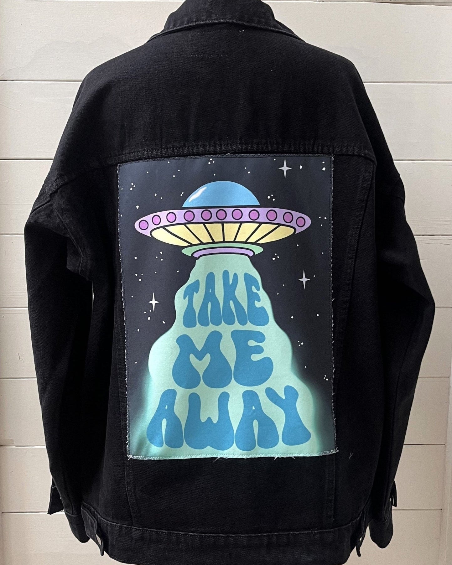 Black jacket with a UFO design and 'Take Me Away' text on the back, displayed against a white wall.