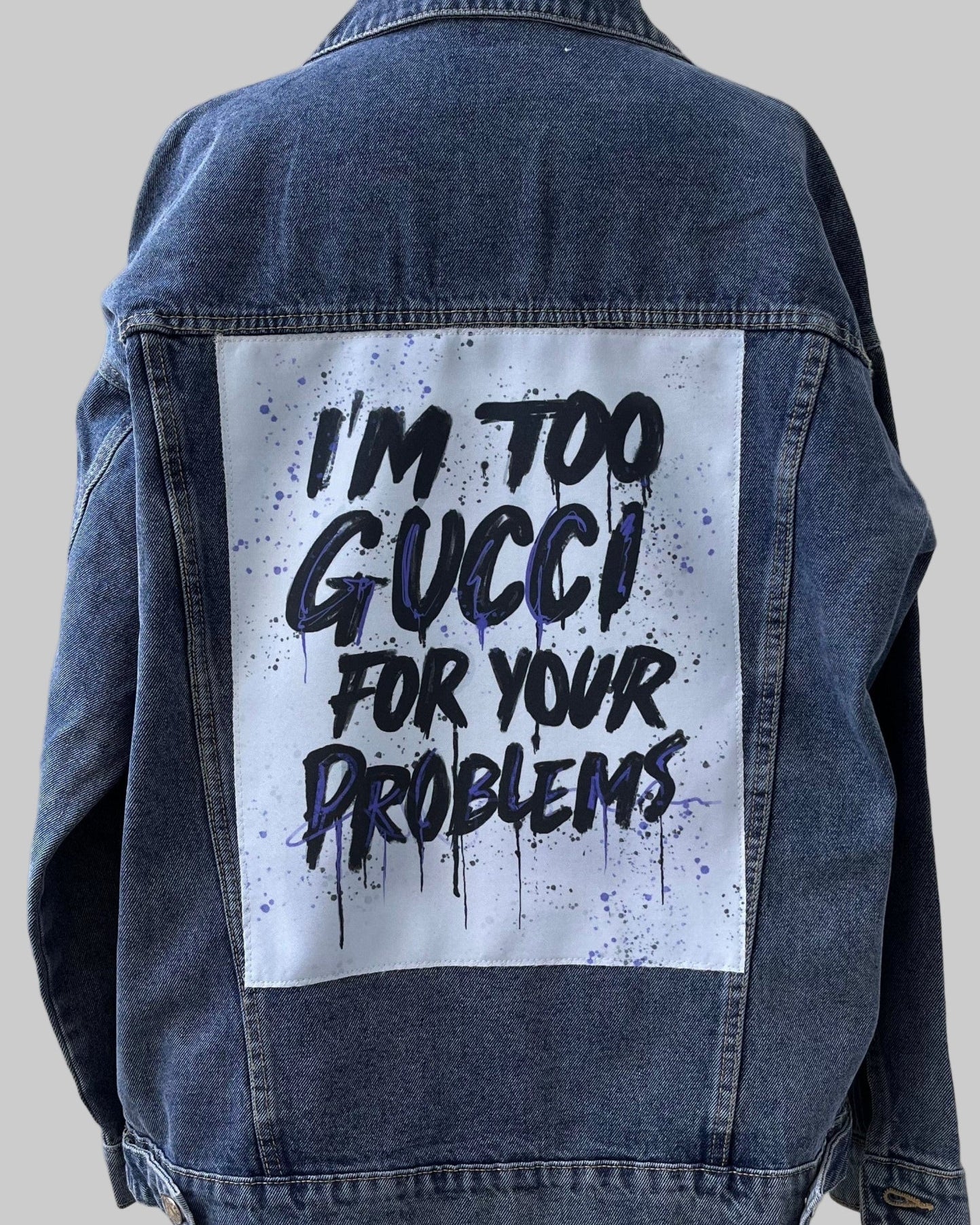 Denim jacket with 'I'm too Gucci for your problems' text on a wooden stand against a light wooden panel background
