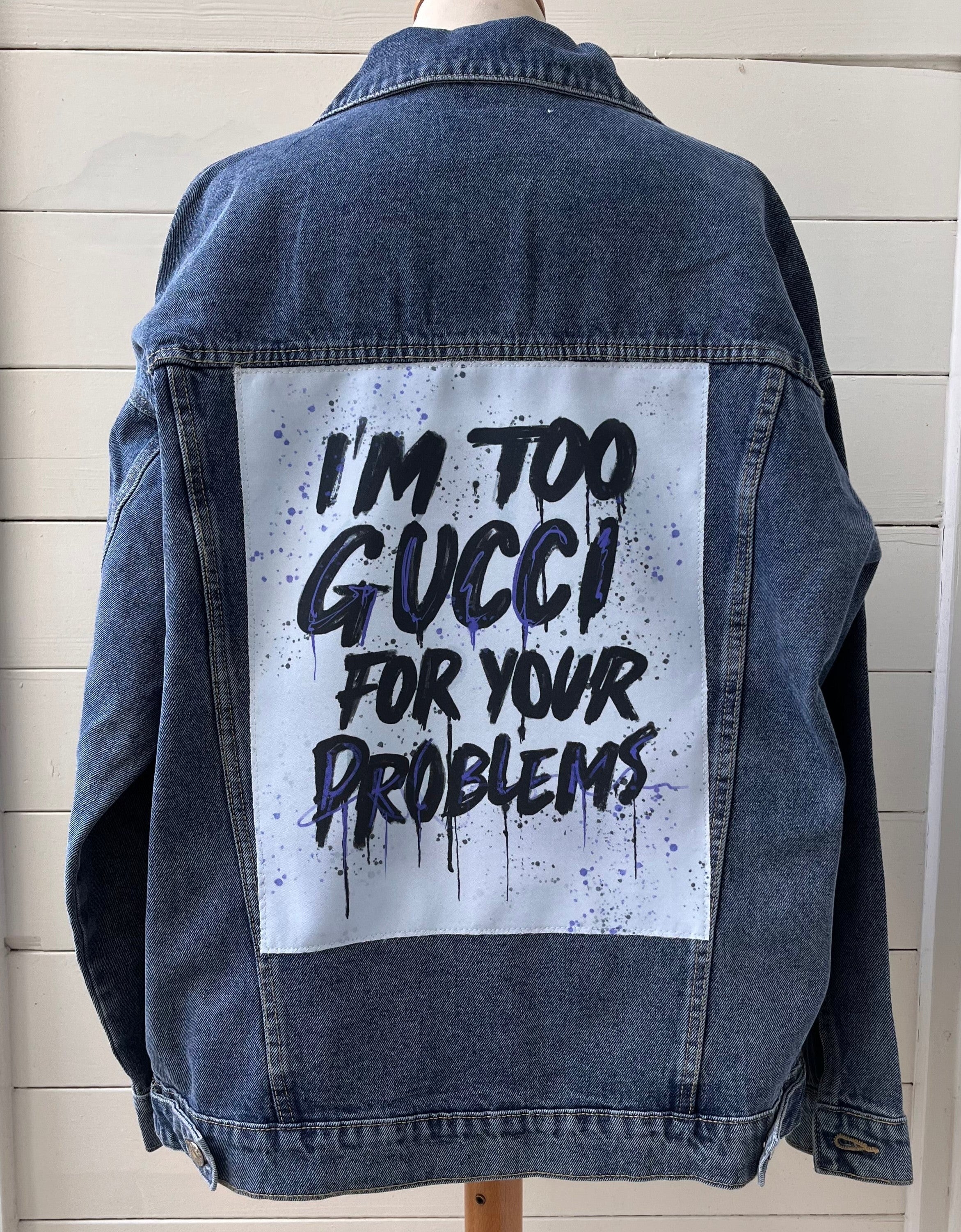 Denim jacket with 'I'm too Gucci for your problems' text on a wooden stand against a light wooden panel background