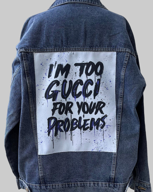 Denim jacket with 'I'm too Gucci for your problems' text on a wooden stand against a light wooden panel background