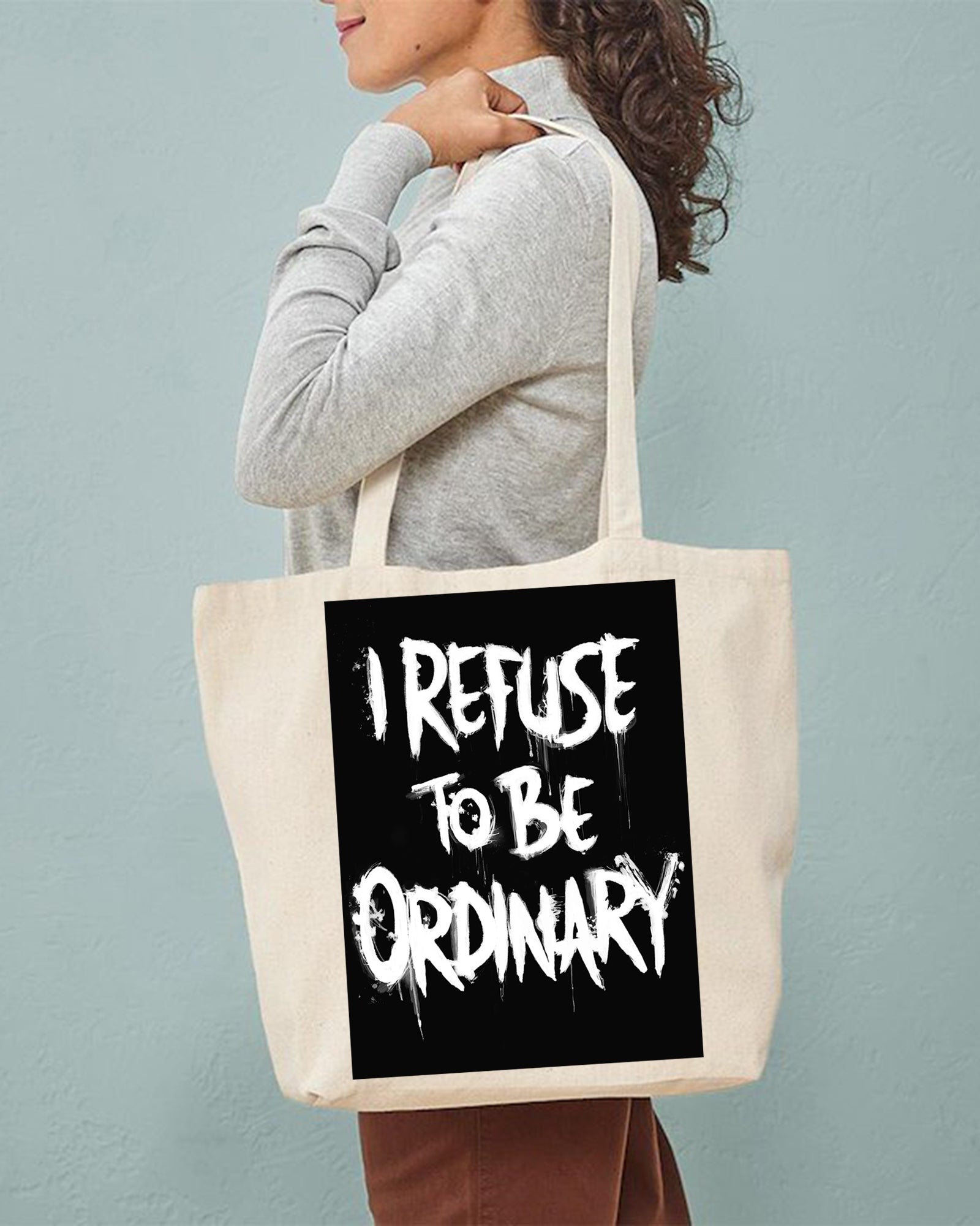 Person holding a tote bag with 'I Refuse to be Ordinary' text against a plain background
