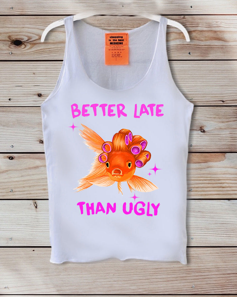 A white sleeveless tank top hanging on a white hanger against a rustic wooden plank background. The tank top features a graphic of an orange goldfish with pink hair rollers and the text 'BETTER LATE THAN UGLY' in pink.