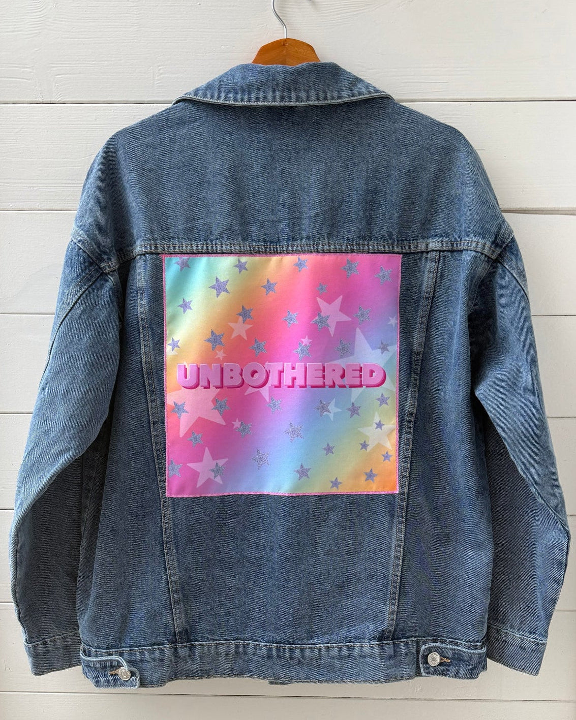 Blue denim jacket with a colorful 'UNBOtherED' graphic on a white wooden background