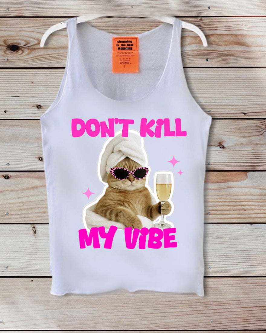 A white tank top displayed on a wooden background, featuring a graphic of an orange tabby cat wearing sunglasses and a white towel, holding a glass of champagne, with the text 'DON'T KILL MY VIBE' in bright pink.