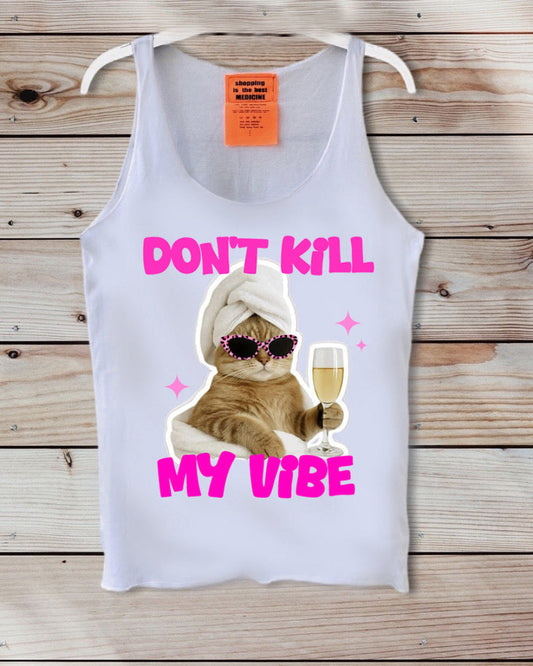 A white tank top displayed on a wooden background, featuring a graphic of an orange tabby cat wearing sunglasses and a white towel, holding a glass of champagne, with the text 'DON'T KILL MY VIBE' in bright pink.