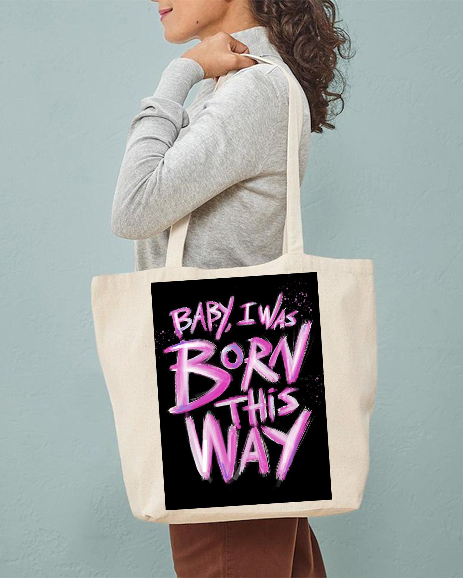 Person holding a tote bag with 'Baby, I was born this way' text on a plain background