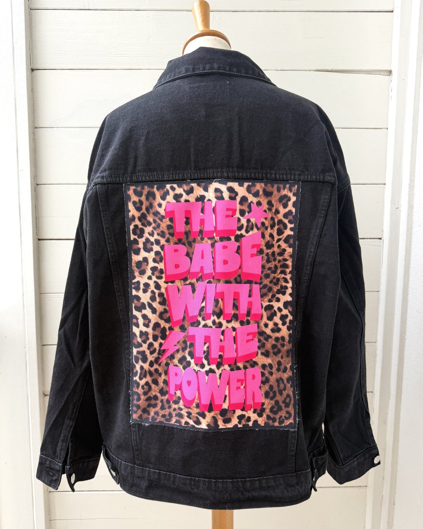 A black denim jacket displayed on a wooden hanger against a white shiplap wall, showing the back panel with a leopard print patch and bright pink text reading 'THE BABE WITH THE POWER'.
