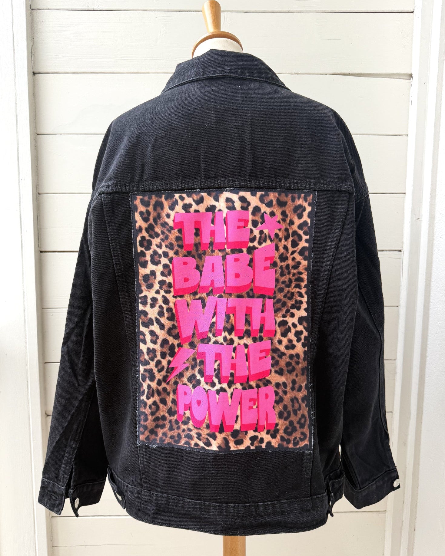 A black denim jacket displayed on a wooden hanger against a white shiplap wall, showing the back panel with a leopard print patch and bright pink text reading 'THE BABE WITH THE POWER'.