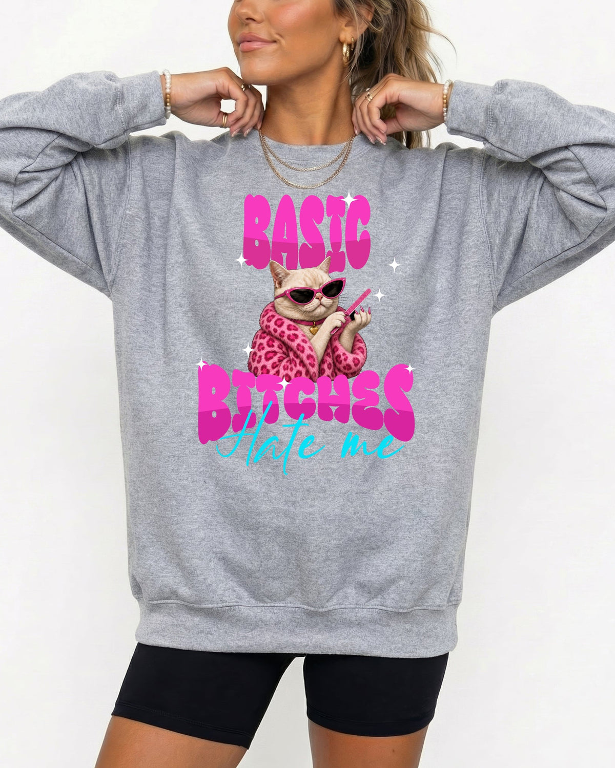 A woman wearing a heather gray sweatshirt with a graphic of a cat in a leopard-print dress and sunglasses, with the text 'BASIC BITCHES Hate me' in pink and blue, paired with black bike shorts against a white background.