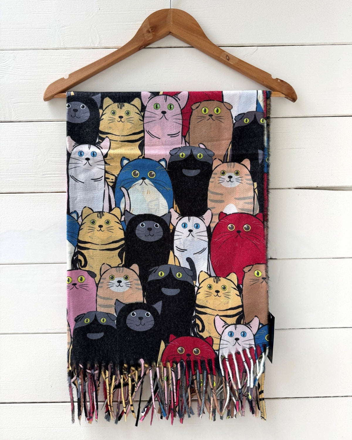 A woven scarf featuring a colorful print of various cartoon-style cats, hanging from a wooden hanger against a white wood-paneled wall.