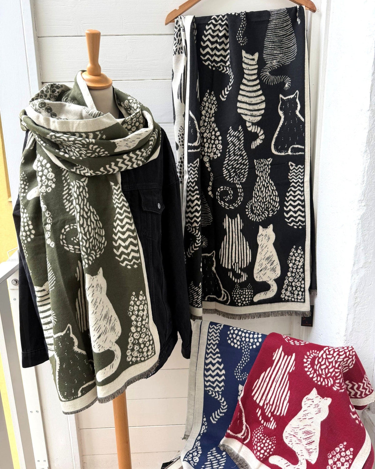 Three woven throw blankets with cat patterns displayed on a wooden hanger and a mannequin torso, with a matching folded blanket on a chair. The blankets feature minimalist cat silhouettes in olive green, black, and navy blue with white, set against a white paneled wall.