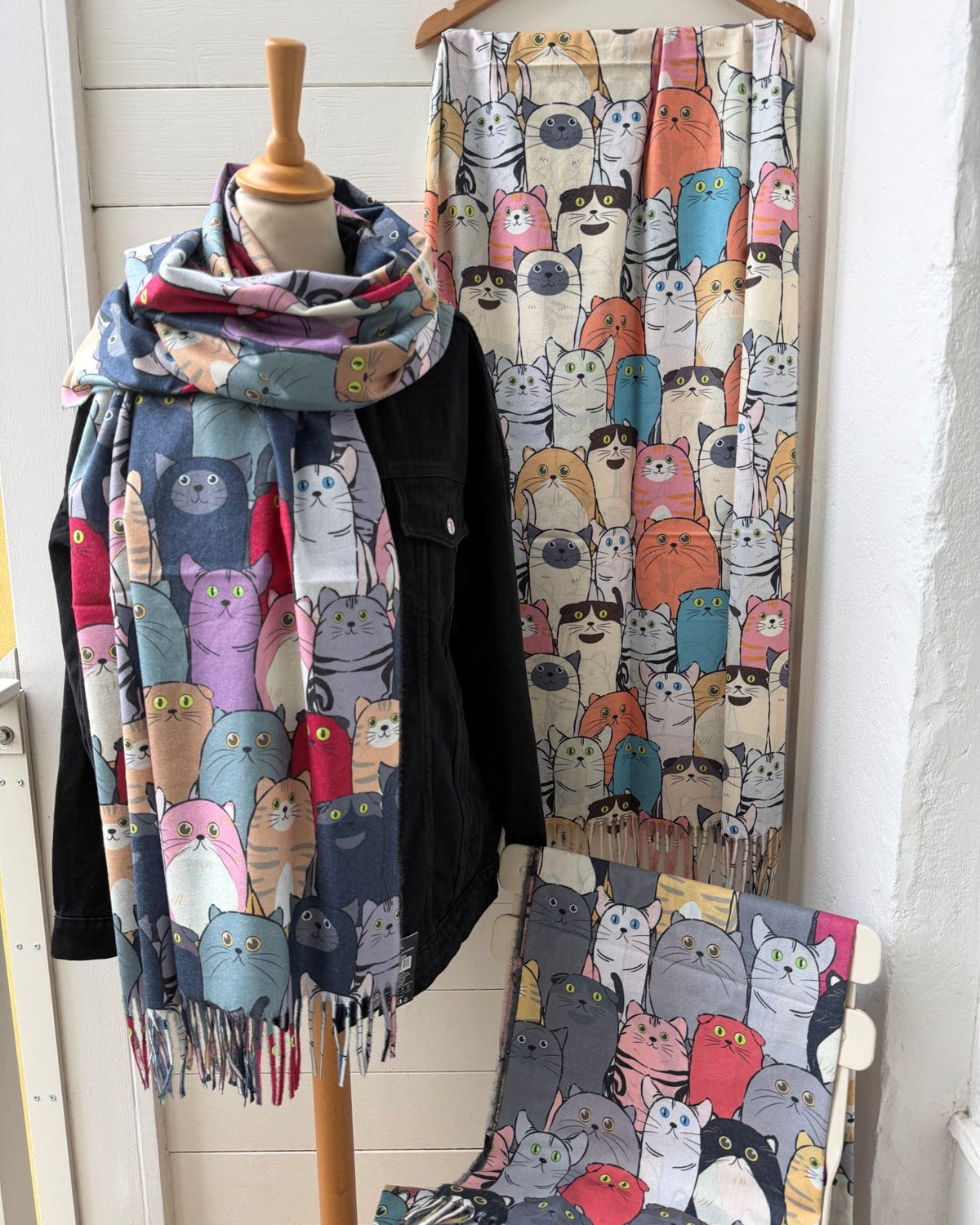 A multicolor scarf featuring a cartoon cat pattern is draped on a wooden mannequin torso, with the same scarf hanging from a wooden hanger on a white paneled wall behind it. A matching chair is partially visible at the bottom right.