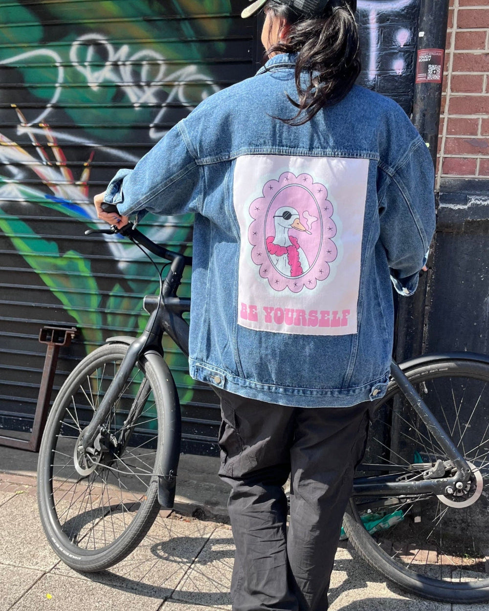 Person wearing a denim jacket with a graphic design and text, standing next to a bicycle against a graffiti-covered wall.