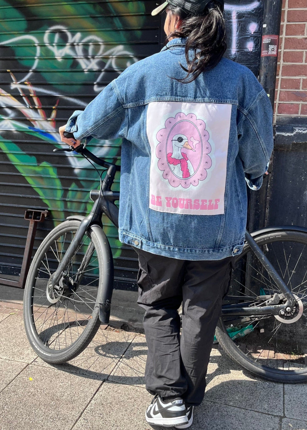 Person wearing a denim jacket with a graphic design and text, standing next to a bicycle against a graffiti-covered wall.