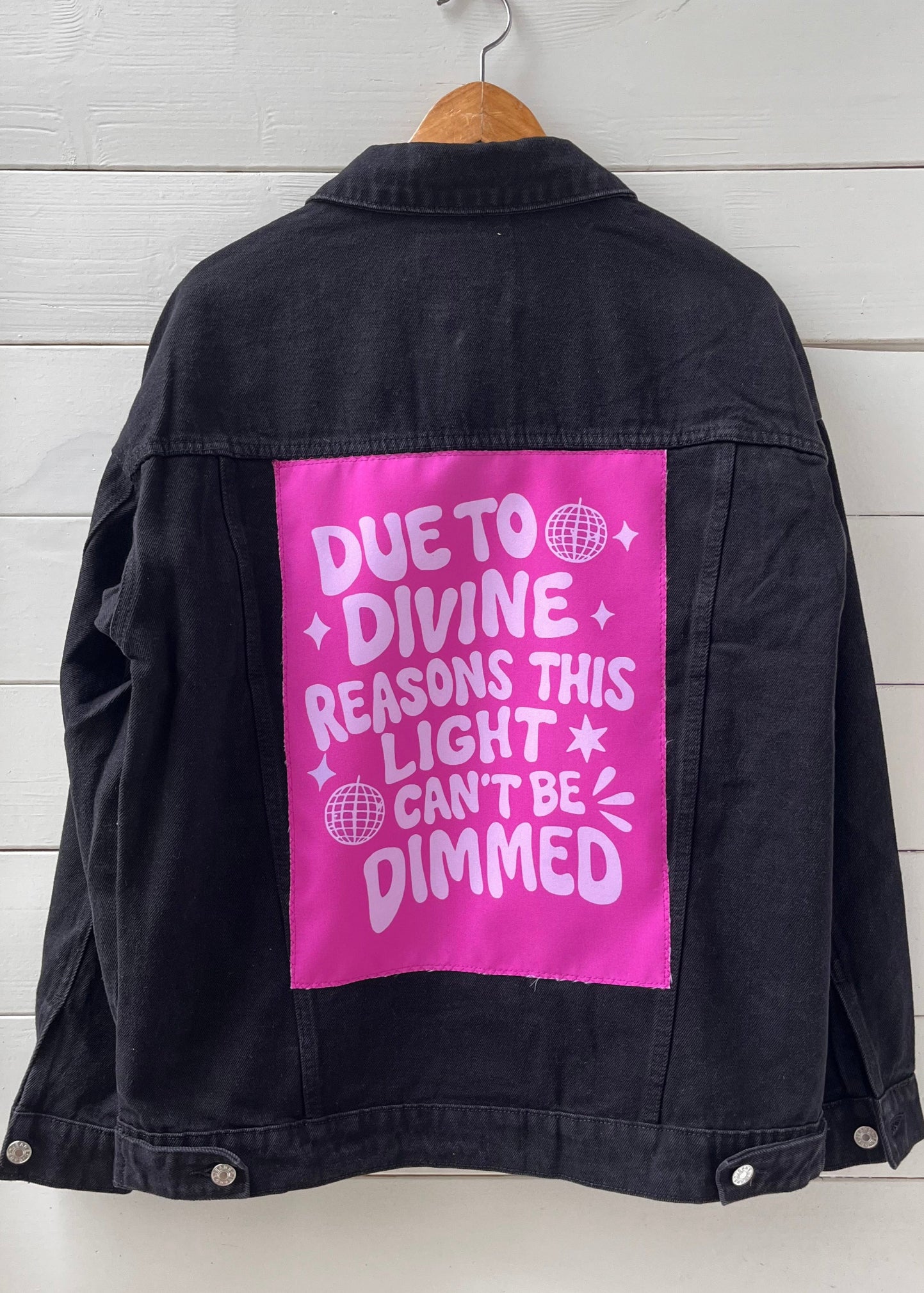 Black jacket with pink patch on a wooden hanger against a white wooden panel background