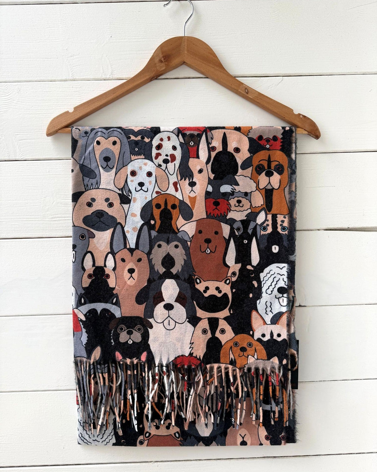 A rectangular scarf with fringe, featuring a dense cartoon illustration of various dog faces on a dark background, hanging from a wooden hanger against a white wood paneled wall.