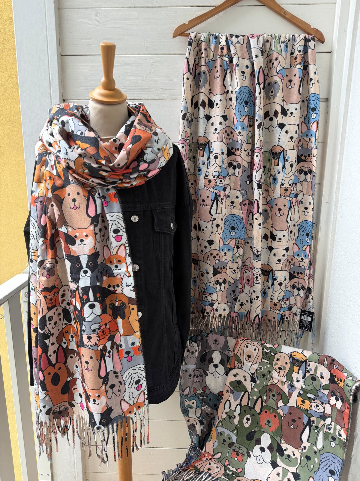 A set of dog-patterned scarves displayed on a mannequin torso and hanger, alongside a matching patterned fabric draped over a chair. The scarves feature various cartoon dog illustrations in multiple colors including black and white, orange, beige, blue, and green.
